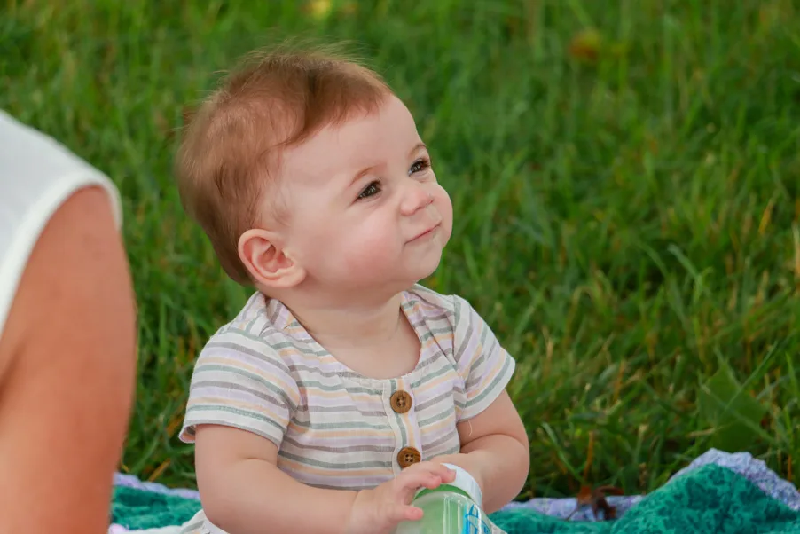 Smiling toddler with red hair wearing a striped shirt sitting on a blanket in green grass outdoors.