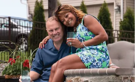 Smiling couple sitting on outdoor patio wall, woman in blue floral dress holding drink, man in navy shirt, greenery in background.