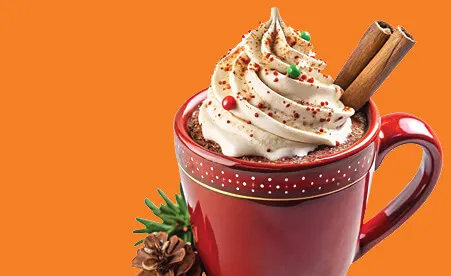 Red mug filled with whipped cream topped hot chocolate, garnished with cinnamon sticks and sprinkles on orange background