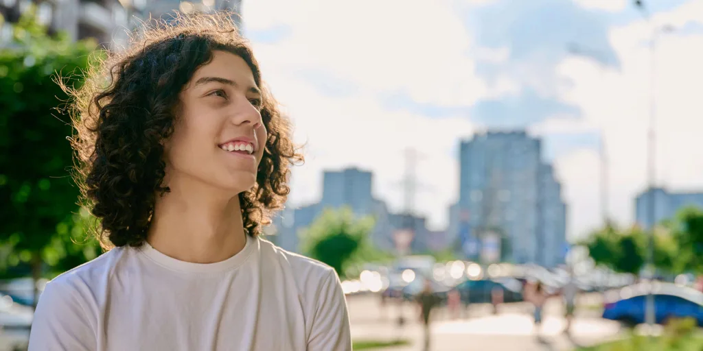 Young man with curly hair smiling outdoors in white shirt with blurred city skyline and greenery in background.