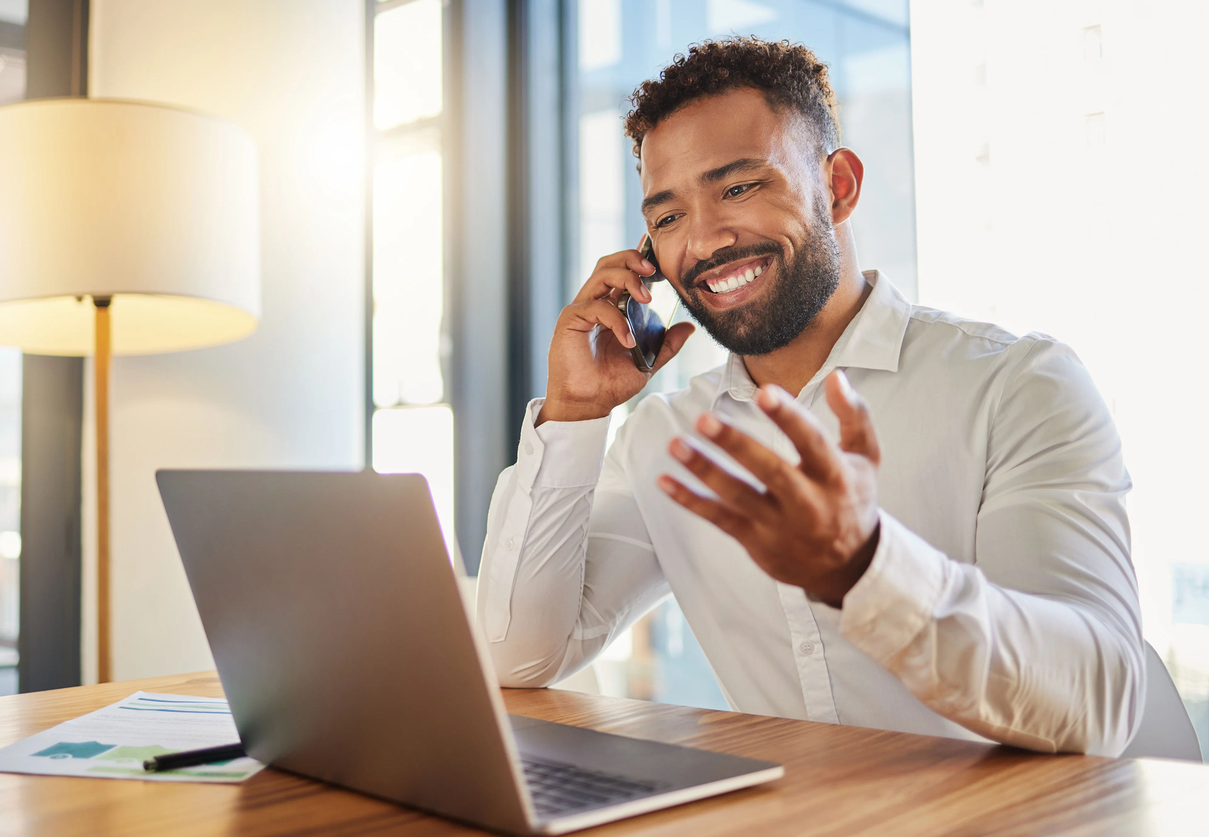 Smiling businessman in white shirt talking on phone while gesturing at desk with laptop in bright modern office.