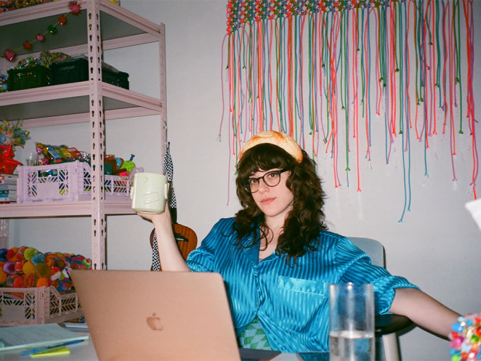 Sam Reece sitting at her workstation holding a mug in one hand, wearing a satin teal shirt and a light orange headband.