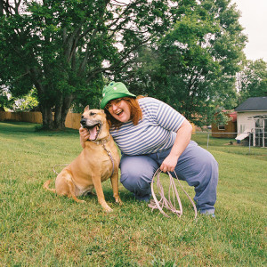 Corook wears a black-and-white striped shirt, jeans, and tennis shoes, paired with a green frog hat while they pose with their dog, Cubby outside on the grass.