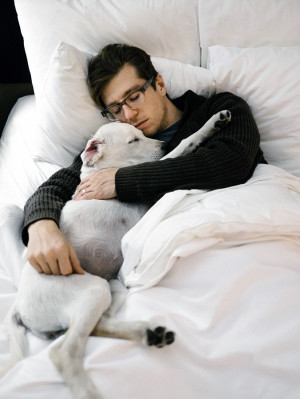 Man in his mid 20's cuddling in bed with his white labrador puppy.

