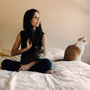 woman sits on a bed looking solemn, alongside her cat