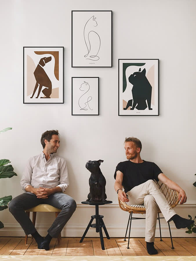 A black dog sitting in between two men with dog and cat illustrations hung on the wall behind them. 