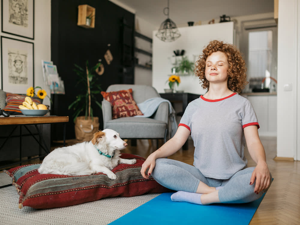 Young ginger haired woman sitting cross legged on the floor in her living room while meditating with her white dog seated on a pillow next to her