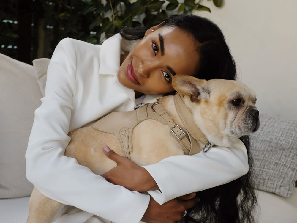 A woman in a long sleeve white shirt hugging a tan pug dog in a beige harness