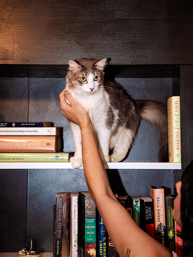 Aparna Brielle reaches her arm up to Oscar Wilde, who rests on a bookshelf. 