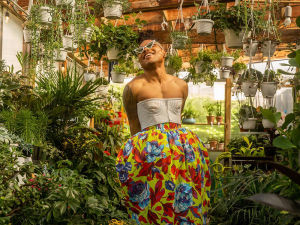 Plant Kween standing in a greenhouse space wearing brightly colored pants and a beige corset top smiling in a carefree way