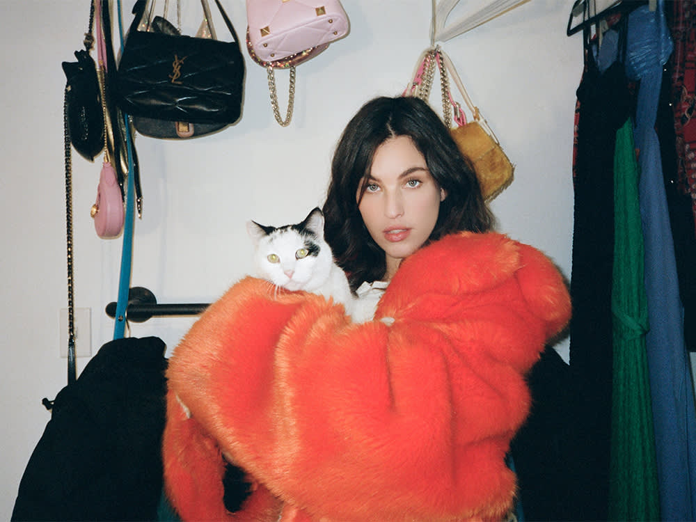 The Wildest's January Cover Star, Rainey Qualley, wearing a bright orange fluffy jacket while holding her white and black cat and standing in a closet full of purses and coats