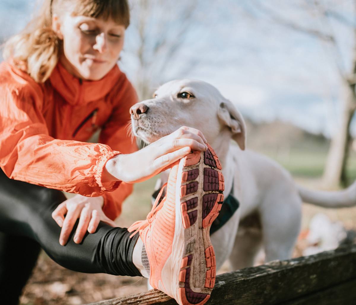 Woman stretching her leg on a park bend with her white dog