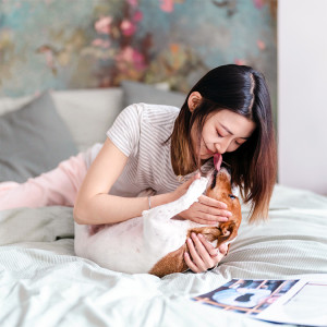 Woman lying on bed with dog, dog is looking up at the woman and licking her in the face
