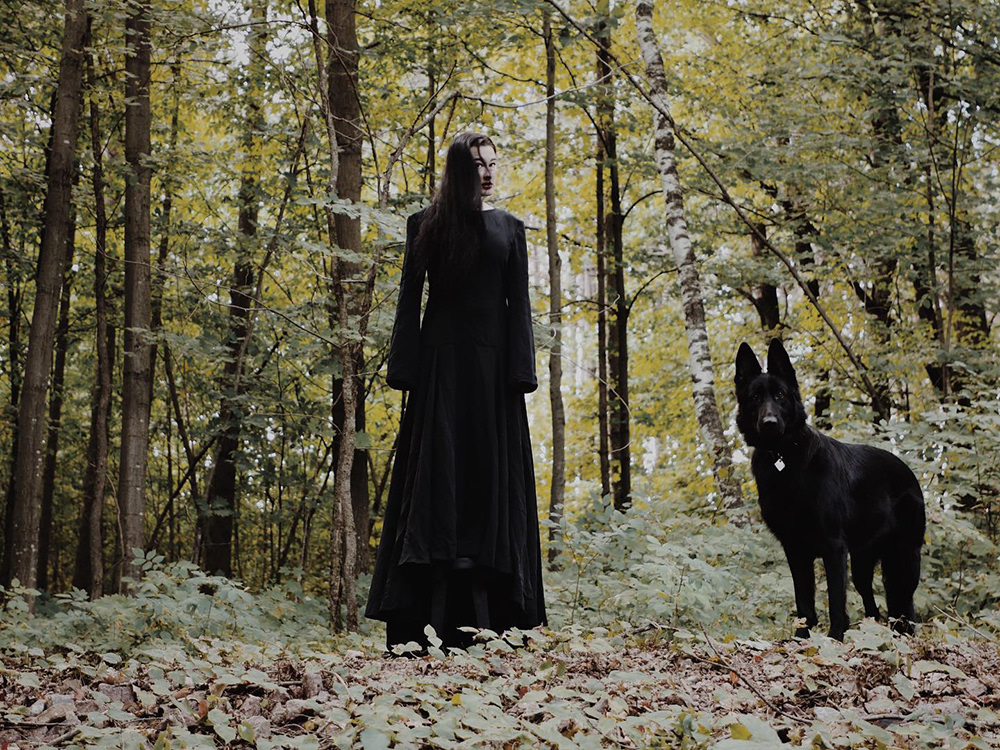 Pet parent and musician Zola Jesus and with her black dog Nadja standing in a dim forest wearing a long black dress
