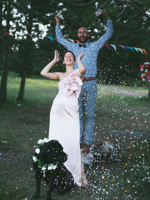 A woman and a man dressed up and celebrating with their arms in the air with confetti falling around them and a dog wearing a flower crown around its neck. 