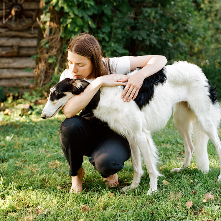 Young woman embracing Greyhound while sitting on green grass and looking away on background of rural house.
