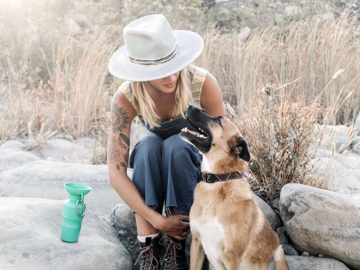 A tattooed blonde woman wearing a white hat looking at her black and tan dog while a Springer pet water bottle sits on a rock behind her on the trail outdoors