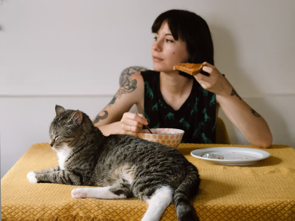A woman eating peanut butter toast with her cat laying on the table