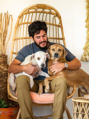 Henry holding two mixed breed dogs in his lap with his arms around them, while sitting in a wicker chair
