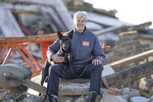 A woman  with cropped grey hair hugs a black dog. They are surrounded by rubble