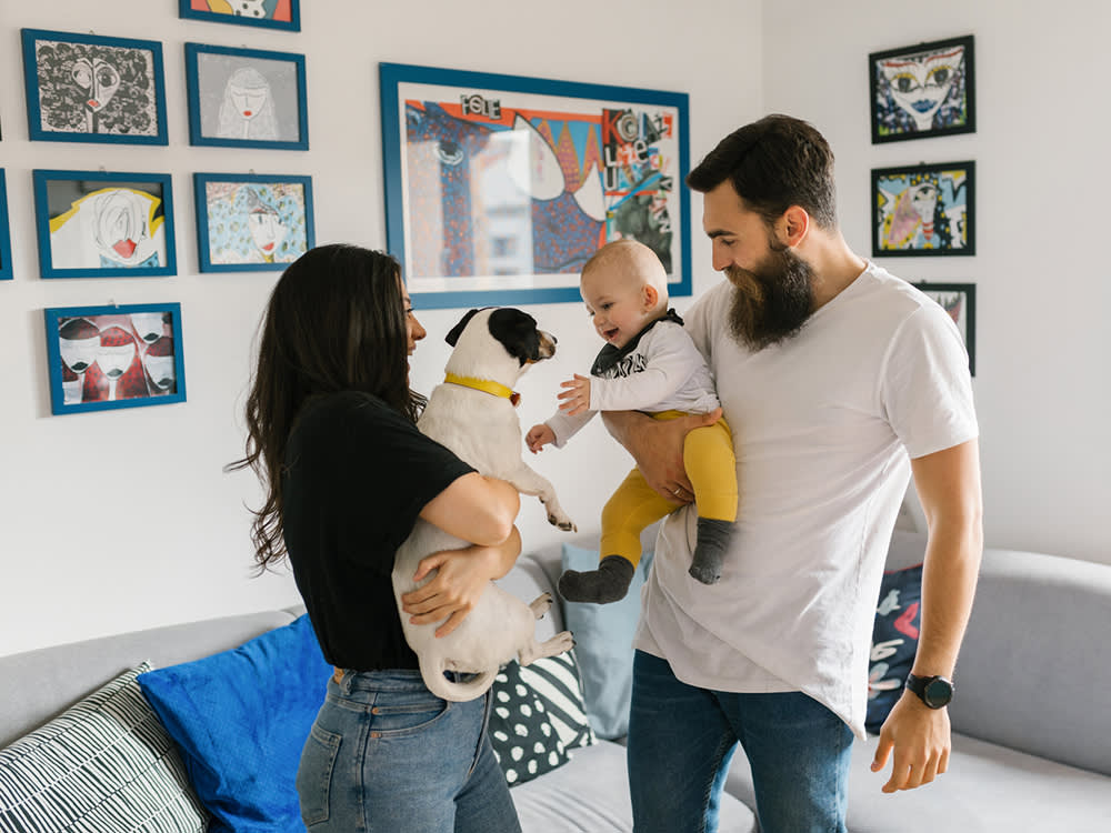 A woman holding her dog and introducing it to a baby in the arms of a bearded man in a white t-shirt and jeans, in the living room