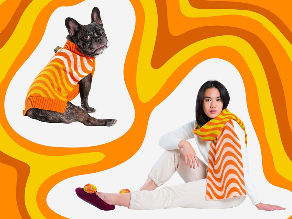 A woman in all white wearing a orange white and yellow striped scarf with a photo of a dark brown Pug dog wearing a sweater that matches the design of the woman's scarf