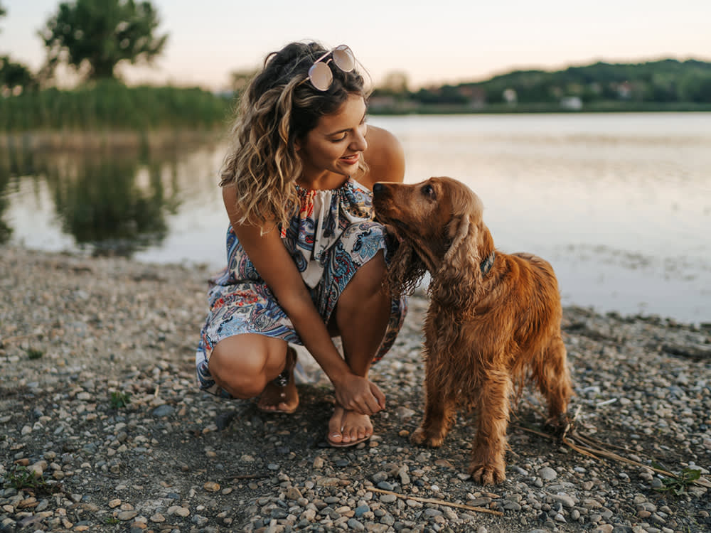 A woman kneeling on the shore of a lake looking at her brown dog standing next to her