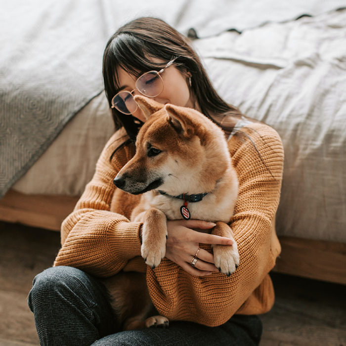 A woman wearing a knit sweater holding a dog close on the floor of her bedroom. 