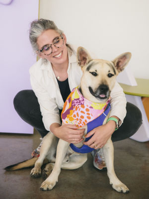 A cheerful woman with a puppy next to her wearing a vibrant harness.