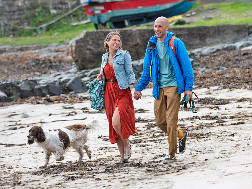 A couple holding hands walk on a UK beach with their dog.