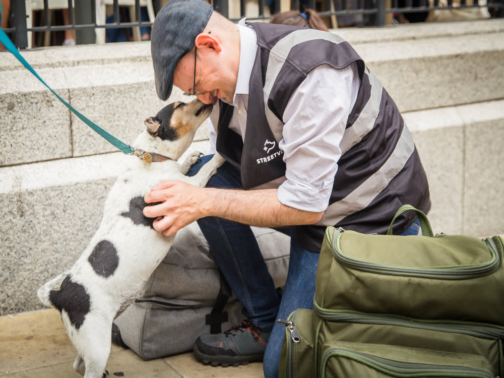 A Streetvet volunteer kneeling on the floor having his face licked by a small dog.