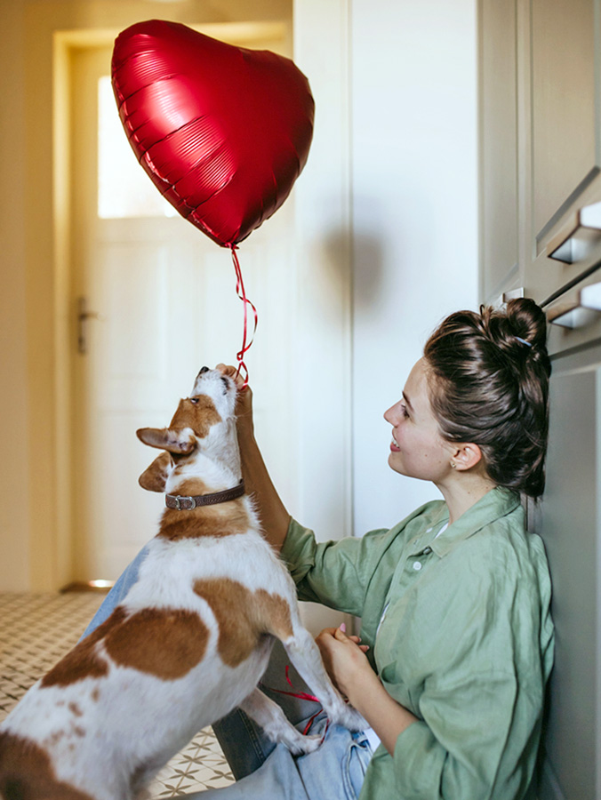 Woman holding a heart balloon with her dog nearby.