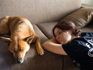 A woman sitting on a floor resting her head on the edge of a couch watching her dog sleep nearby. 
