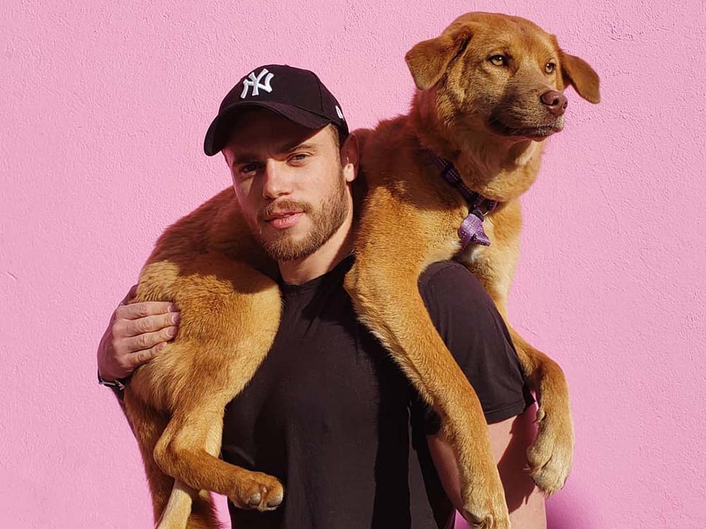 Gus holding his dog Birdie on his shoulders against a solid pink background