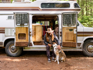 A man with long brown hair and a wide brimmed tan hat sitting in the open part of a van while petting his mixed breed dog in the woods