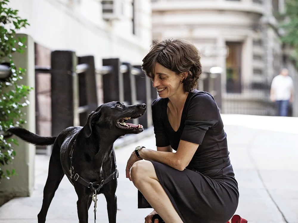 Alexandra Horowitz kneeling outside next to a happy black dog with a smile. 