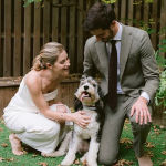 A bride and groom kneeling on their backyard lawn petting their happy dog