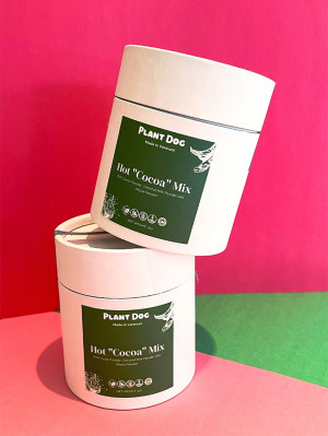 Two jars of Plant Dog's Hot "Cocoa" Mix for dogs stacked in front of a pink and green paper background