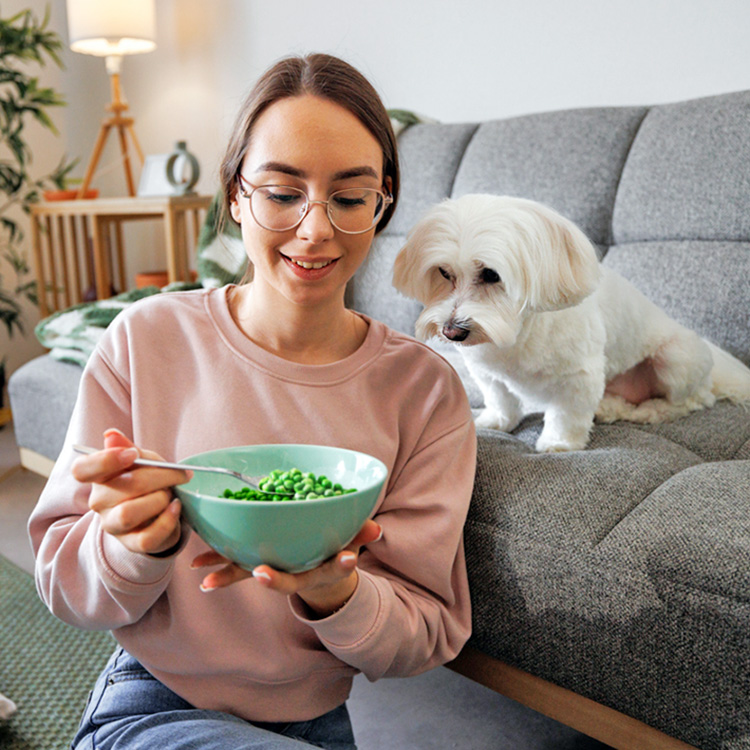 Woman eating peas in a bowl in front of her small white dog.