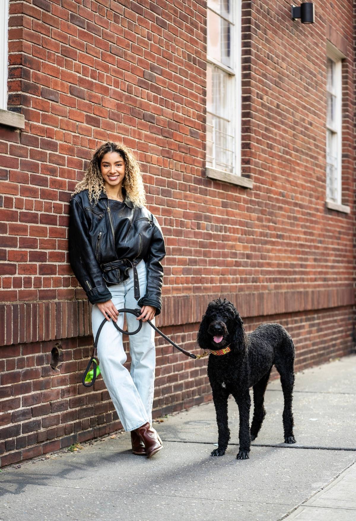 a picture of a cool woman with curly hair leaning against a brick wall and holding the lead of black poodle