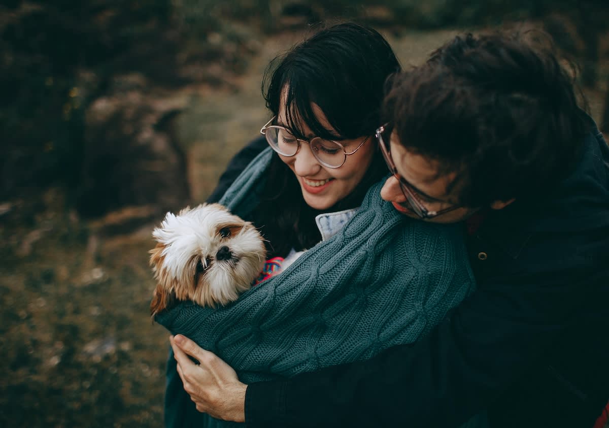 a dark haired couple with glasses holds a tiny dog wrapped in a scarf
