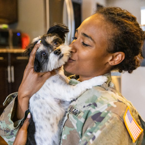 A woman in a U.S. Army uniform hugging a small dog in her arms in the kitchen