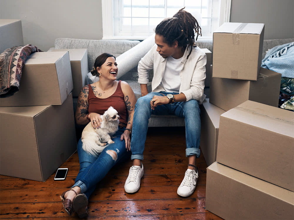 a woman and a man sit in an empty home surrounded by boxes with a dog 