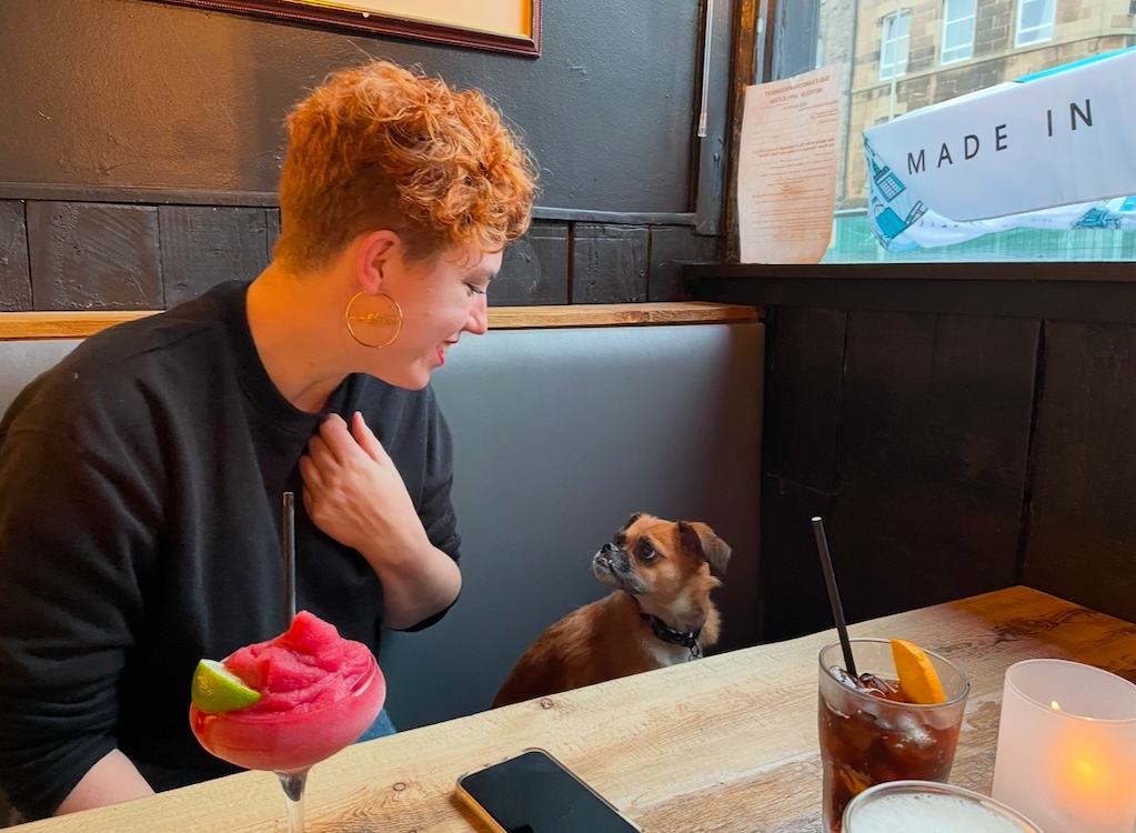 a woman and a dog look at each other in a booth with cocktails