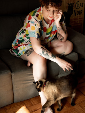a tattooed person with curly red hair on a couch pets a brown and black cat