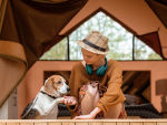 A woman sitting outside of a tent wearing a fedora and headphones shaking a dogs paw. 