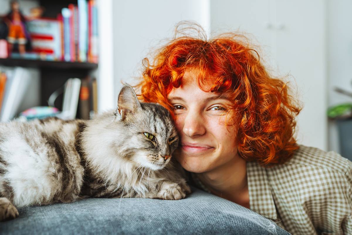 a woman with a curly red bob poses with her face next to a long haired tabby cat