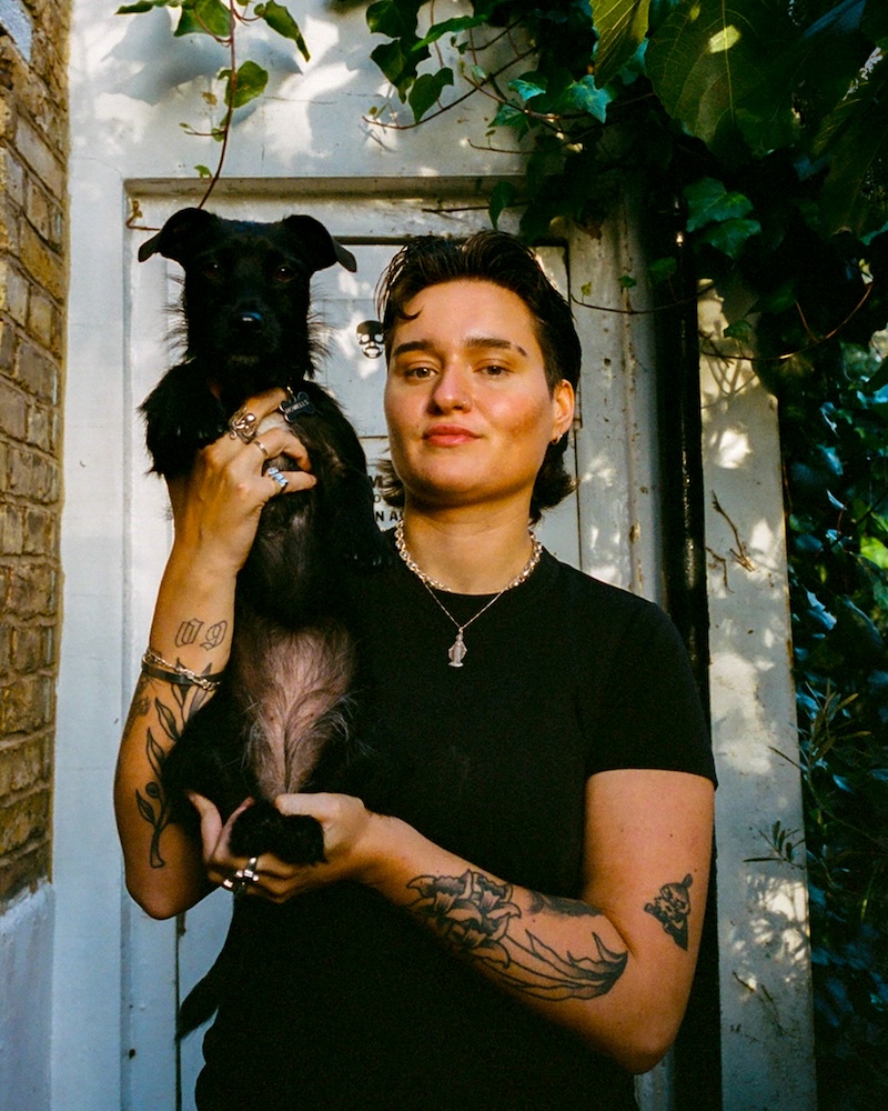 a person with tattoos and black hair holds up a small black dog
