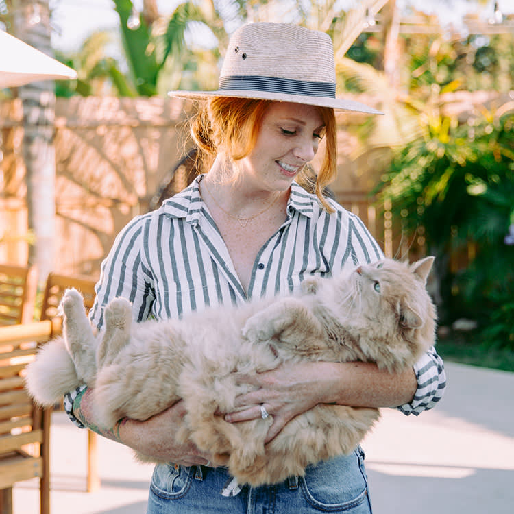 Young woman smiles while standing outside wearing a sunhat and holding up her big fluffy cat.