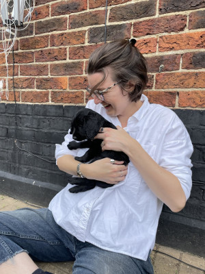 girl with short brown hair and glasses in a white shirt cuddling a small black puppy